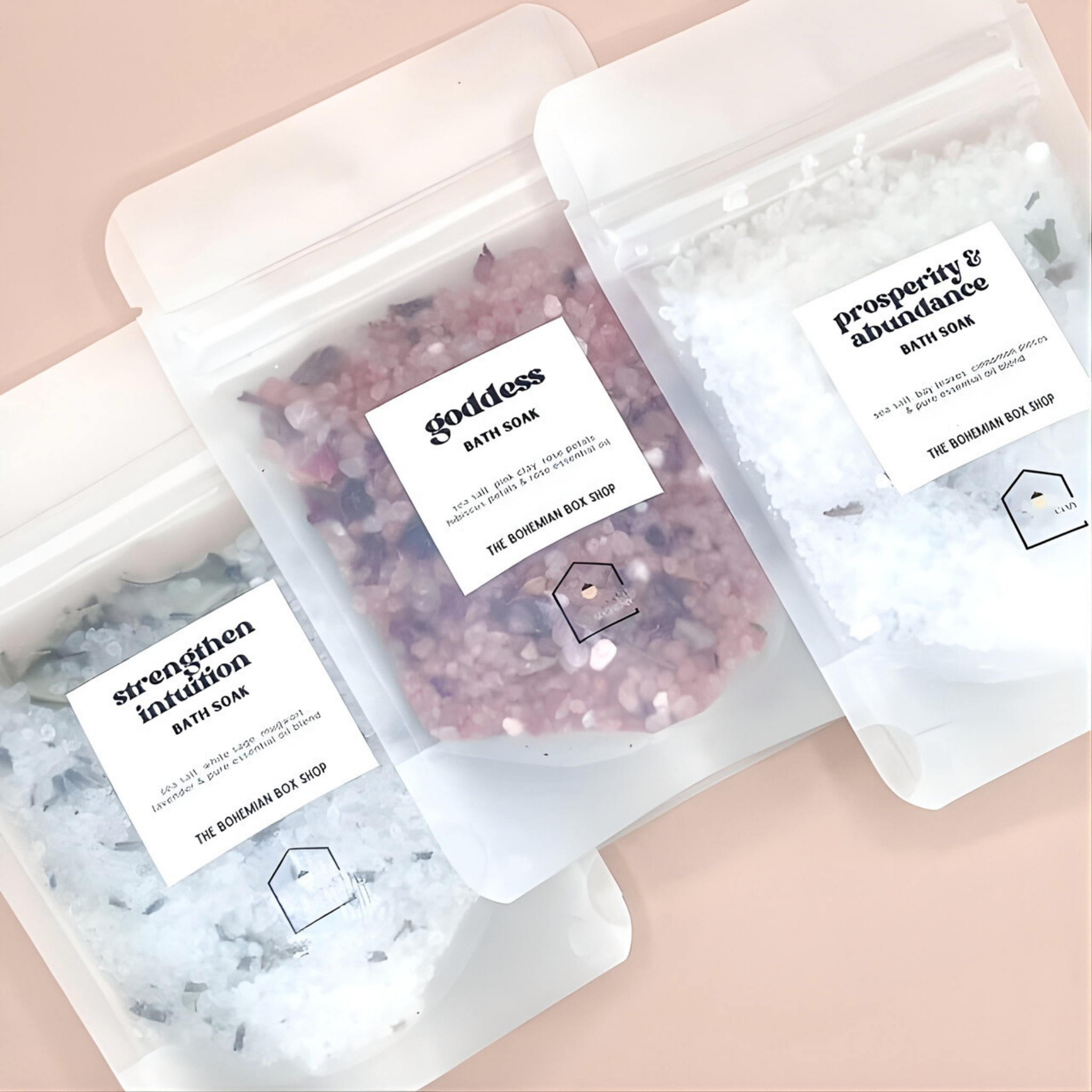 Three bath soaks in clear packaging with labels on a pink background