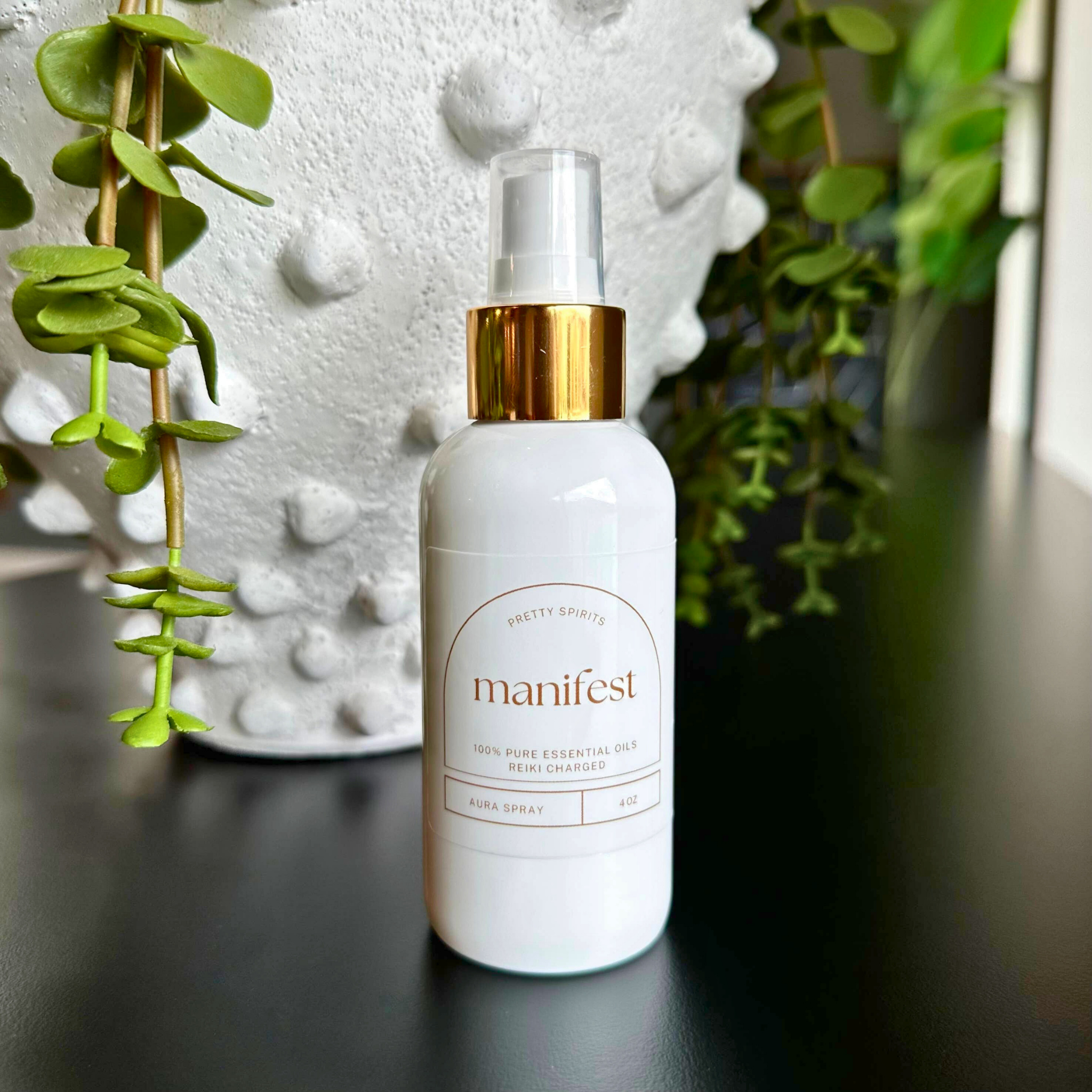 White spray bottle labeled 'manifest' with gold cap on a dark surface with green plants in the background.