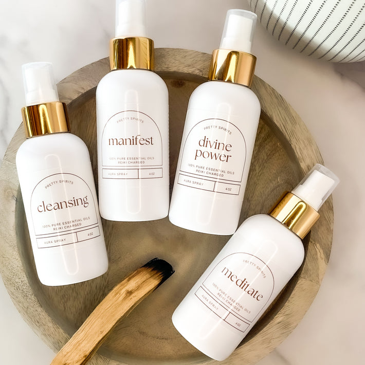 Four white bottles with gold caps on a wooden tray, featuring labels like 'manifest', 'divine power', 'cleansing', and 'meditate'.