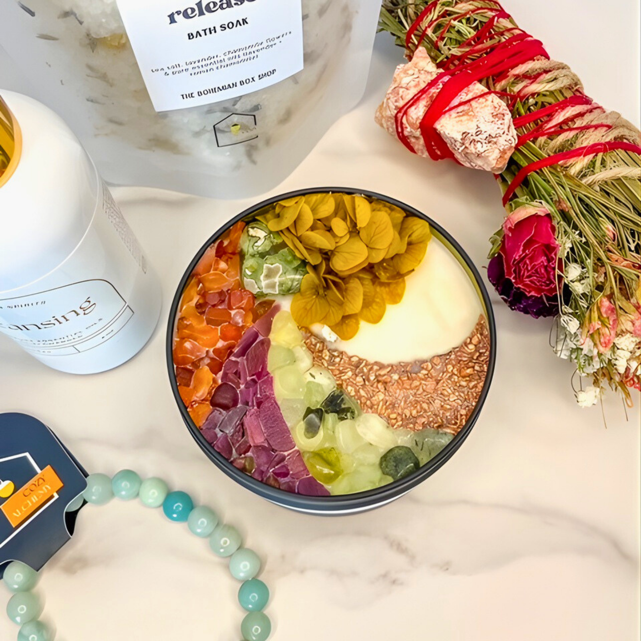 Colorful bath bomb in a black bowl with a candle, flowers, and packaging on a marble surface.