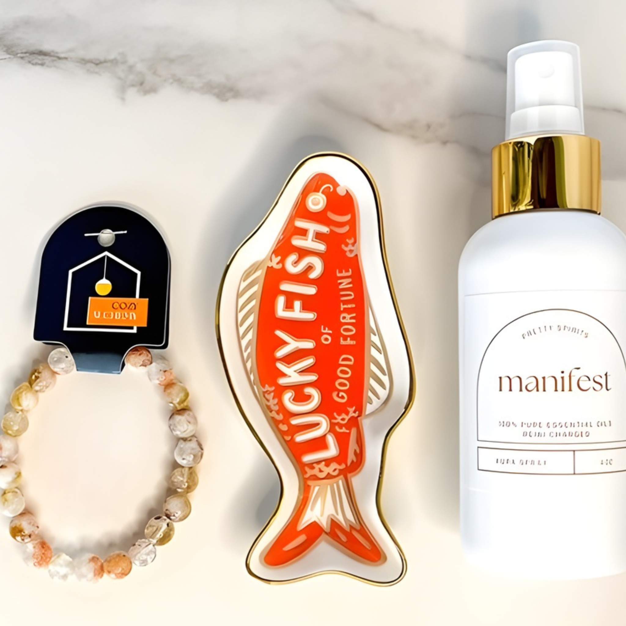 Collection of 'Lucky Fish' good fortune charm, bracelet, and 'manifest' essential oil spray bottle on a marble surface.