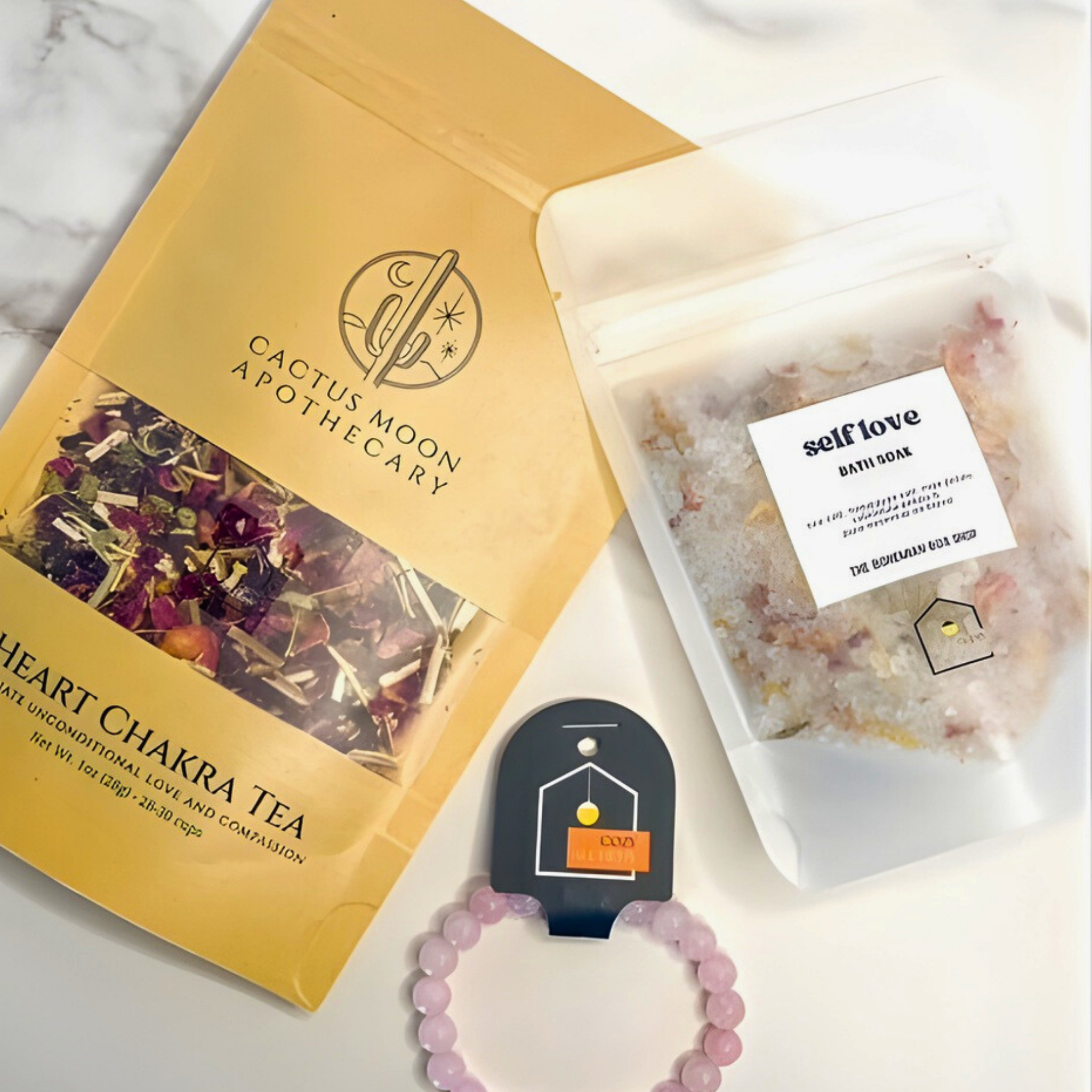 Cactus Moon Apothecary heart chakra tea package, self-love bath soap, and a bracelet on a marble surface.