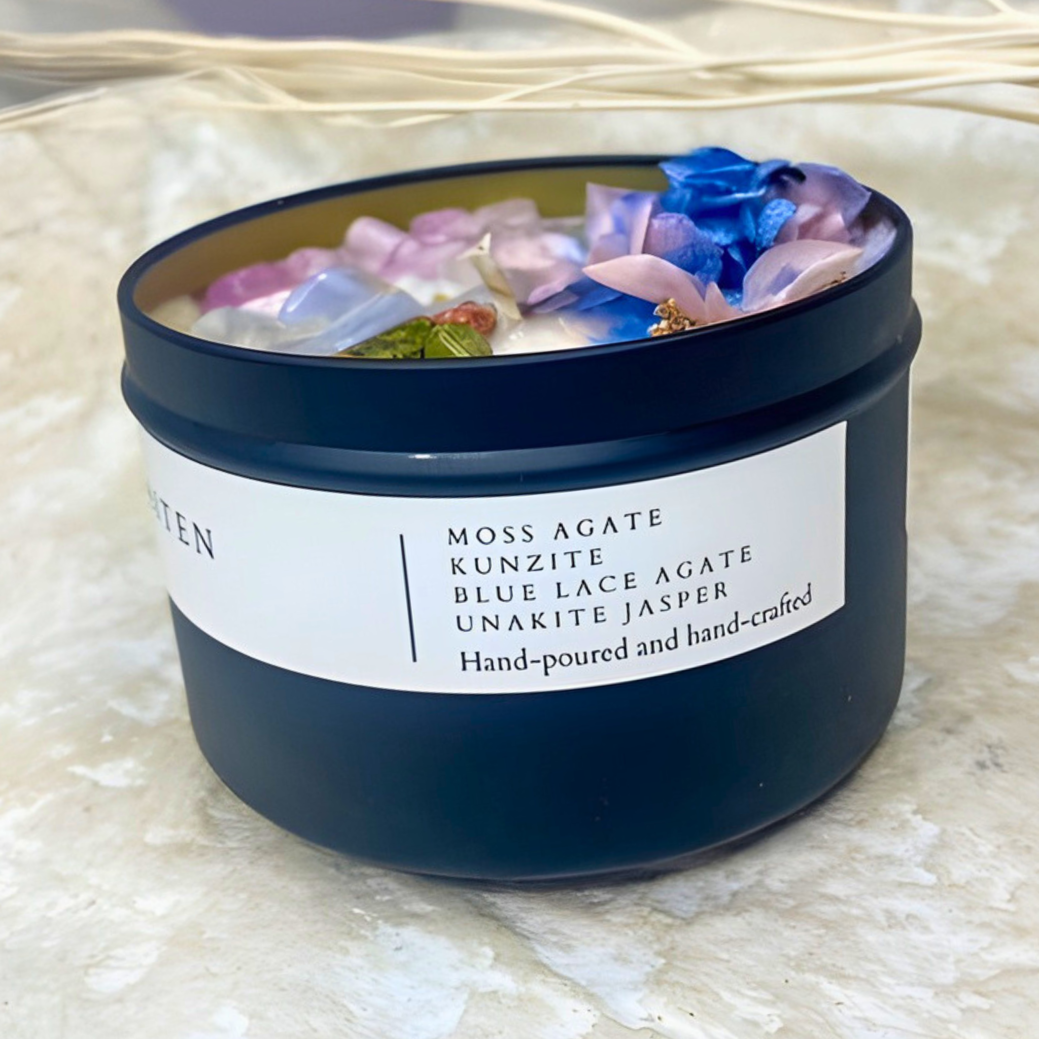 Candle in a black container with decorative stones and flowers on a textured surface