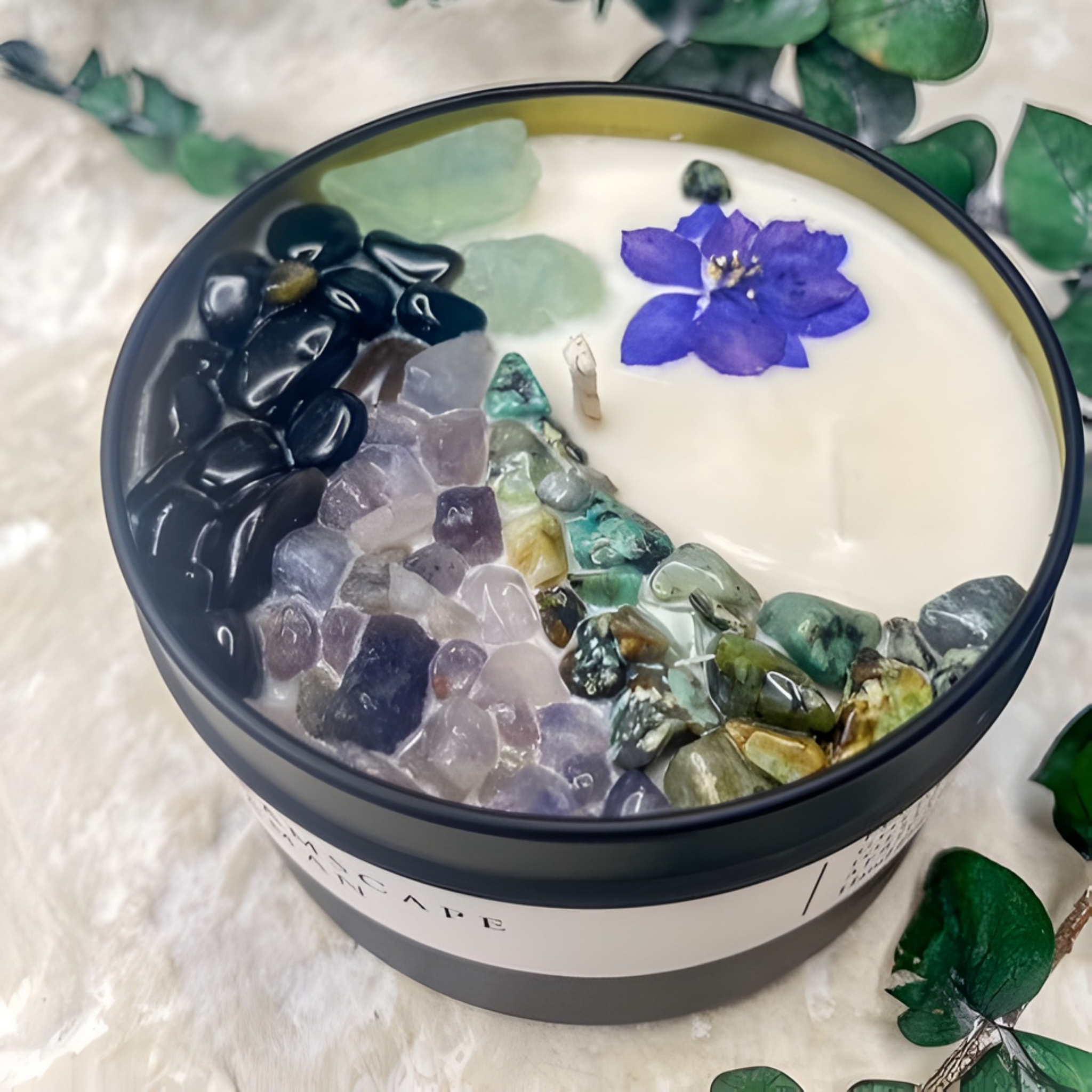 Candle with decorative stones and a purple flower on a textured surface with green leaves.