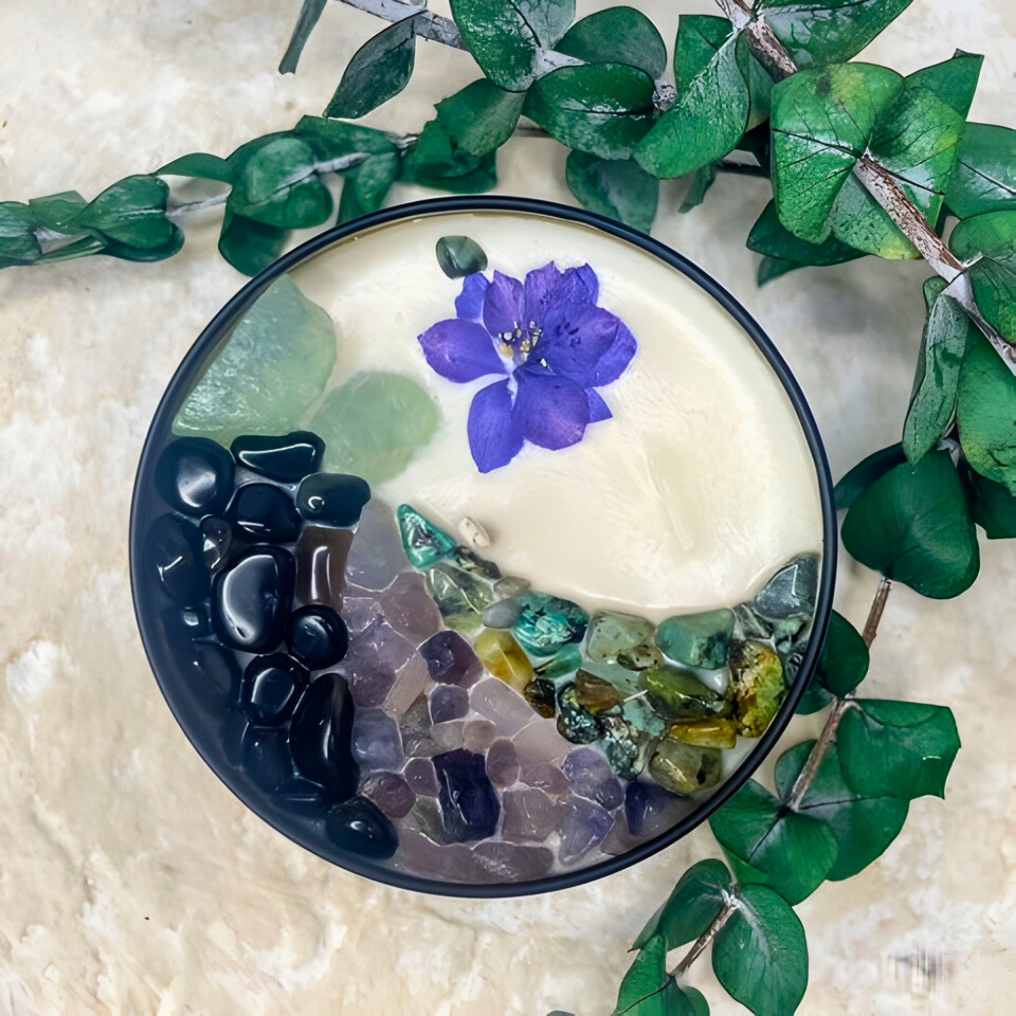 Decorative bowl with stones and a purple flower on a textured surface with green leaves.