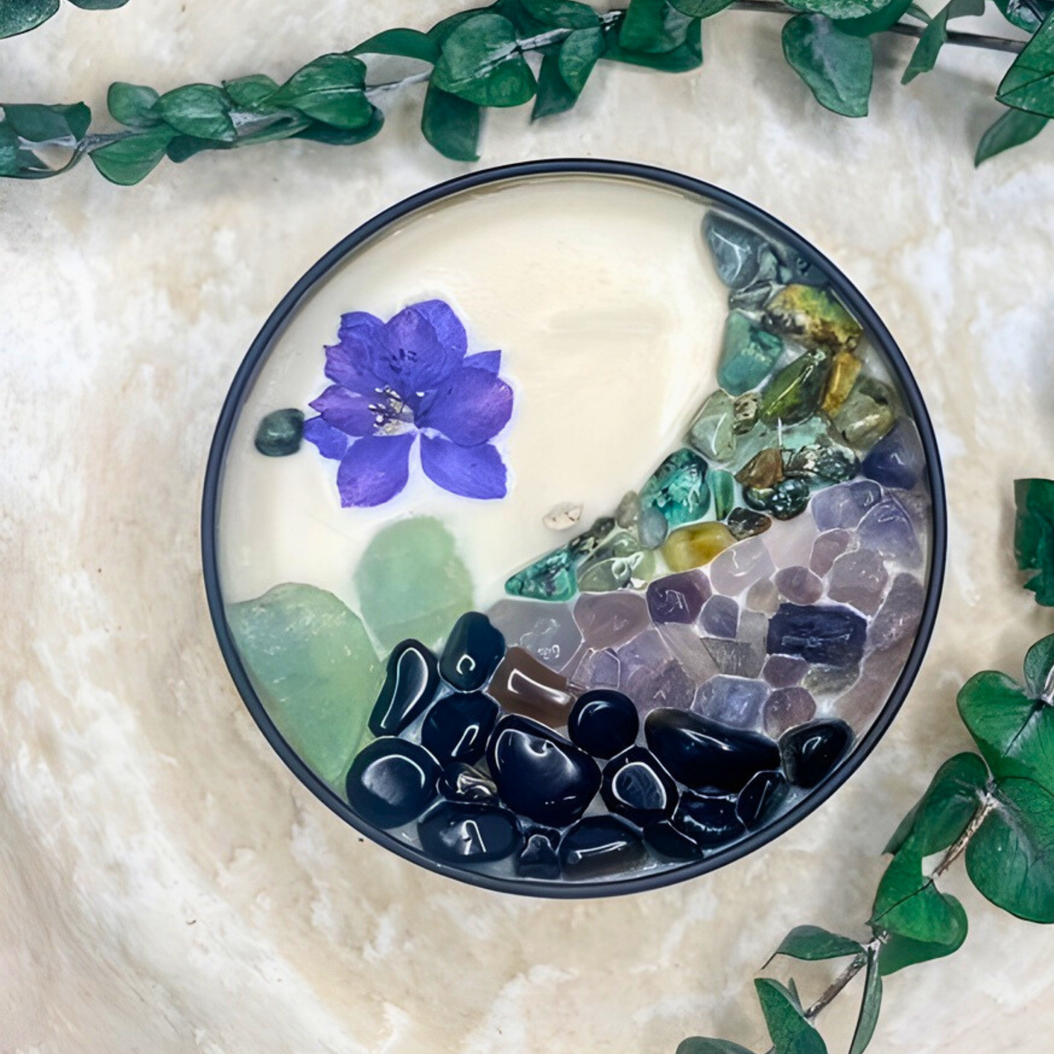Decorative stone bowl with a purple flower and colorful stones on a textured surface with green leaves.