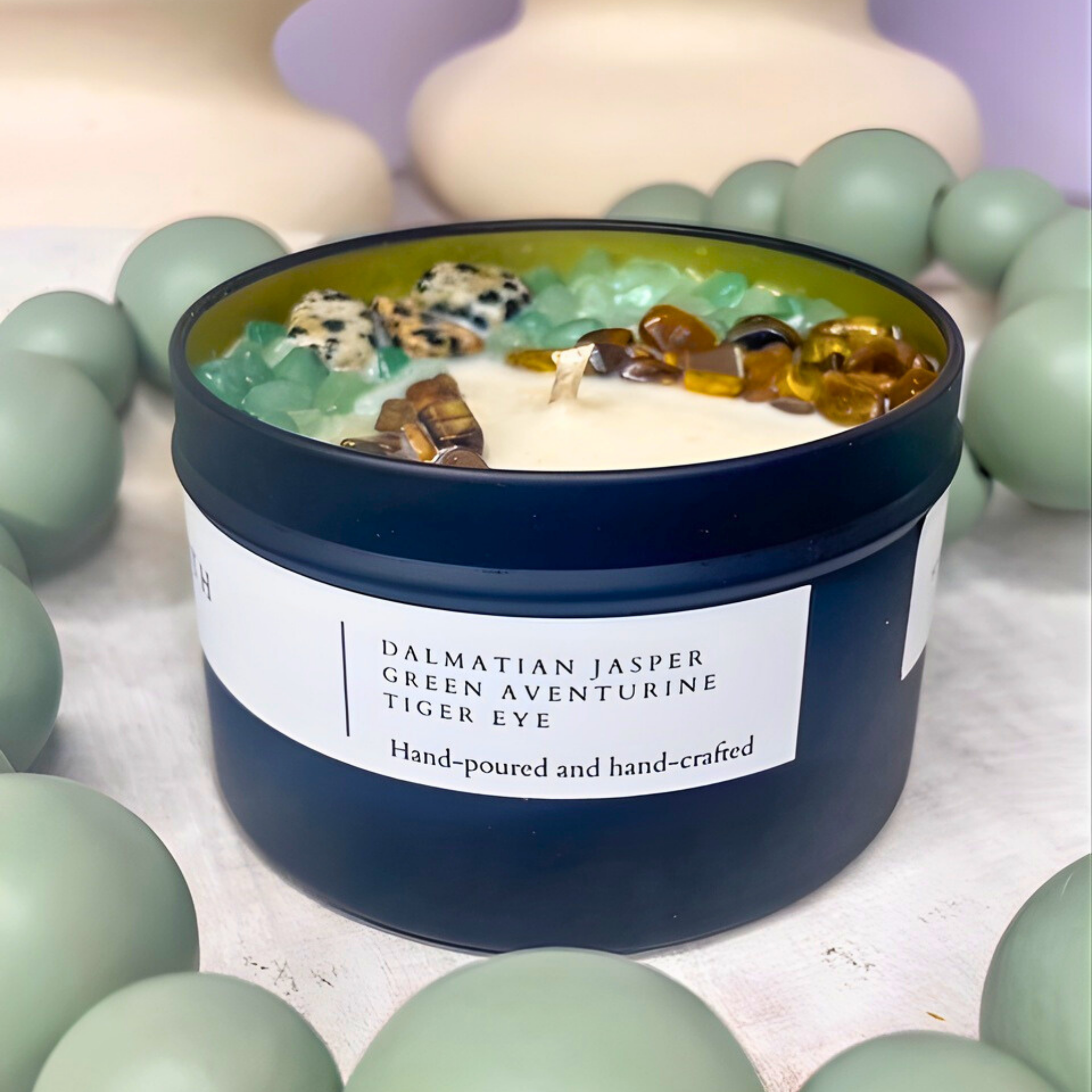 Candle in a blue container with decorative stones, surrounded by green spheres on a light background