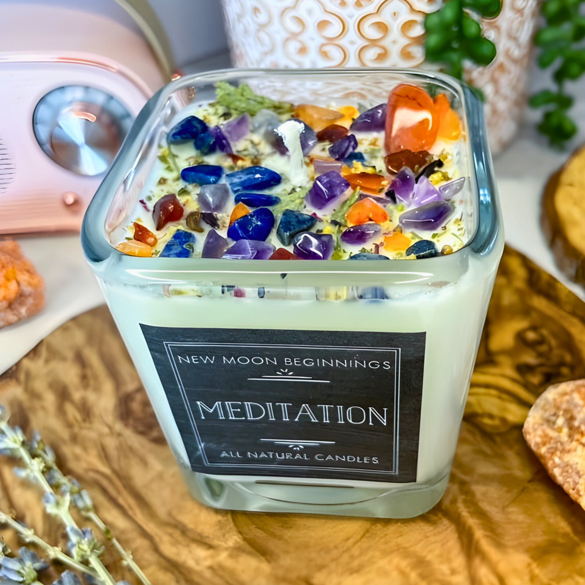 Candle with colorful stones labeled 'New Moon Beginnings Meditation' on a wooden surface.