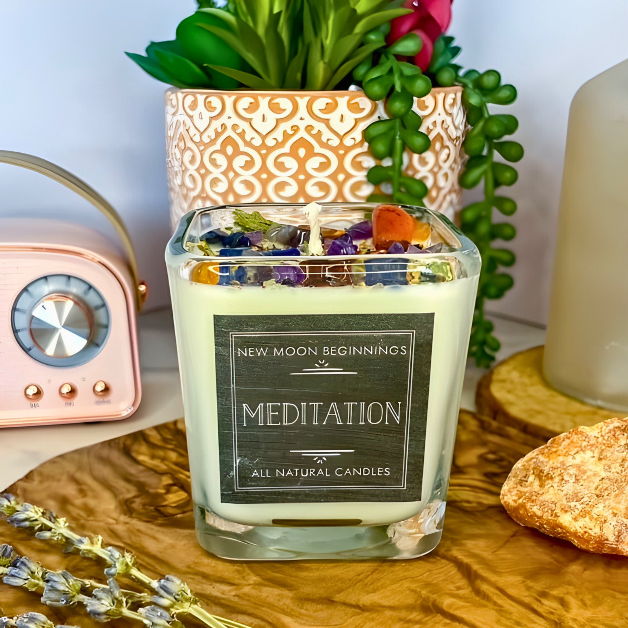 Meditation candle labeled 'New Moon Beginnings' on a wooden surface with plants and a radio in the background.