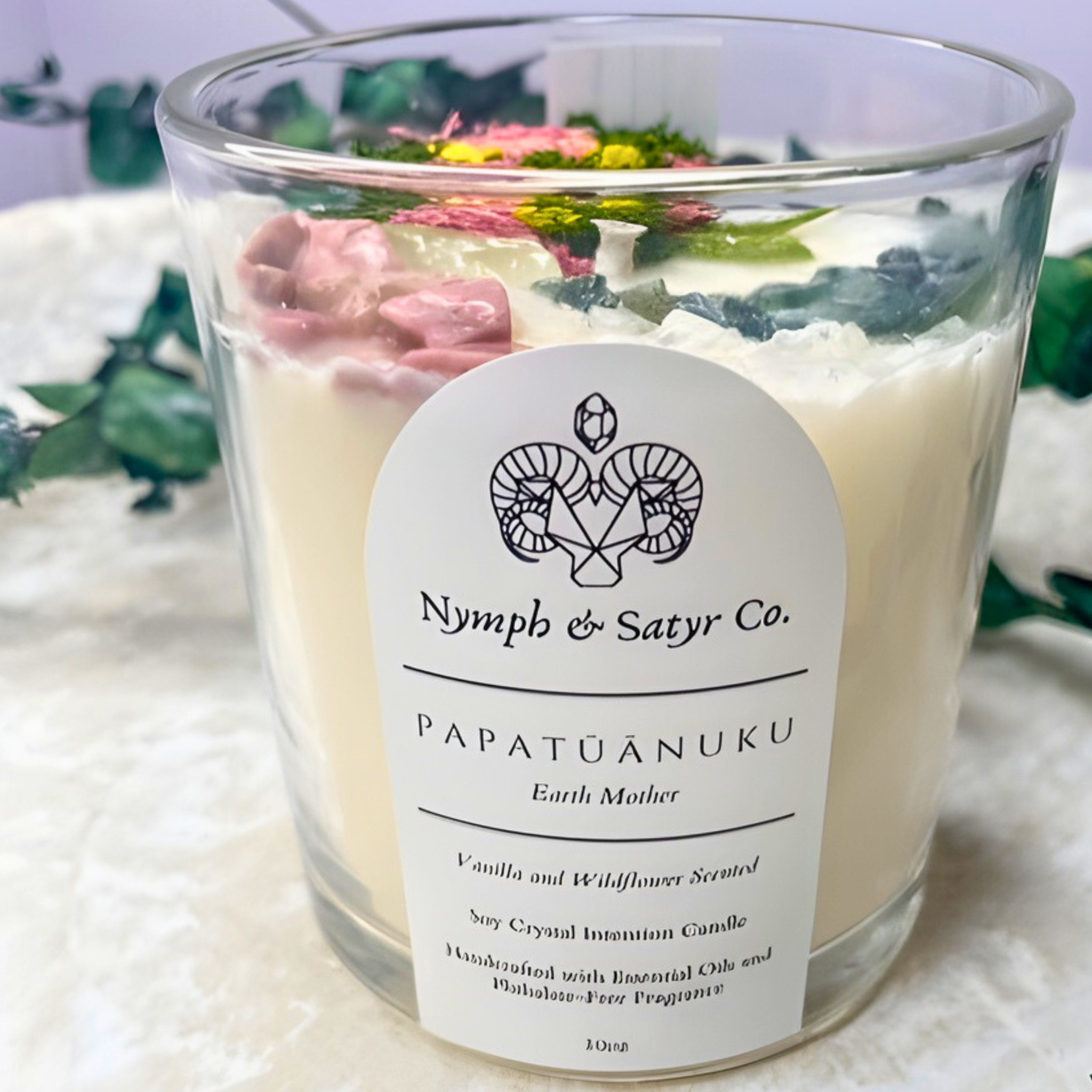 Candle with a label reading 'Nymph & Satyr Co.' on a white surface with greenery in the background.