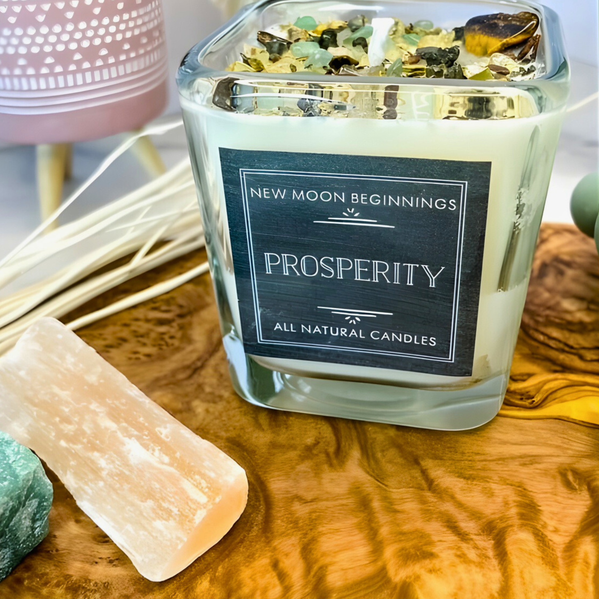 Glass candle labeled 'Prosperity' by 'New Moon Beginnings' on a wooden surface with decorative elements.