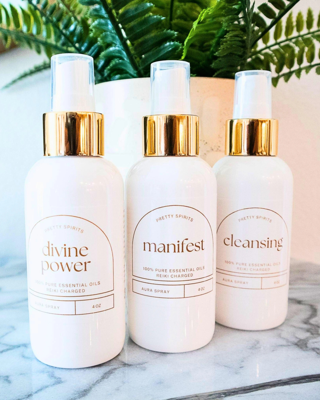 Three bottles of essential oils labeled 'divine power', 'manifest', and 'cleansing' on a marble surface with greenery in the background.