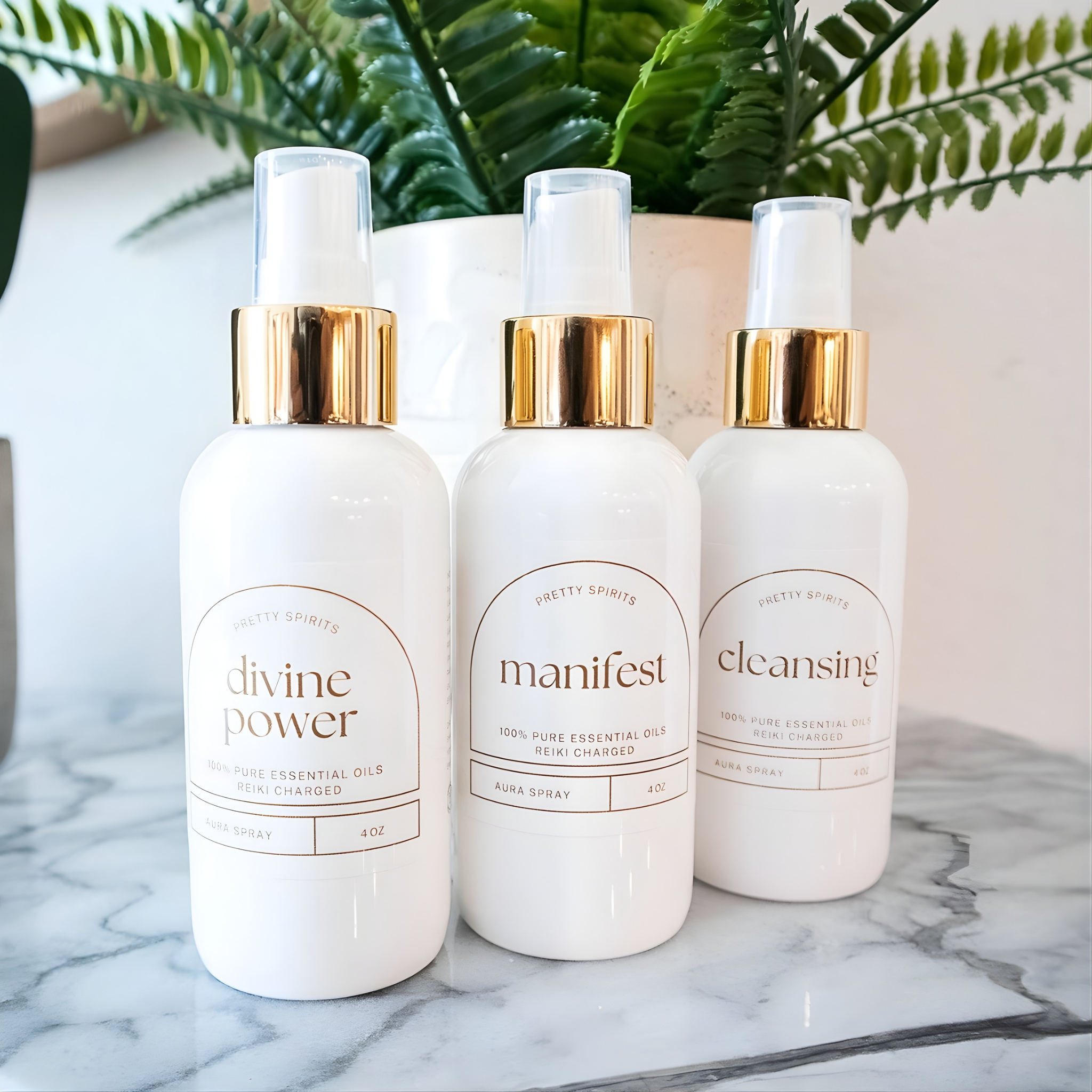 Three white bottles with gold caps labeled 'divine power', 'manifest', and 'cleansing' on a marble surface with a plant in the background.