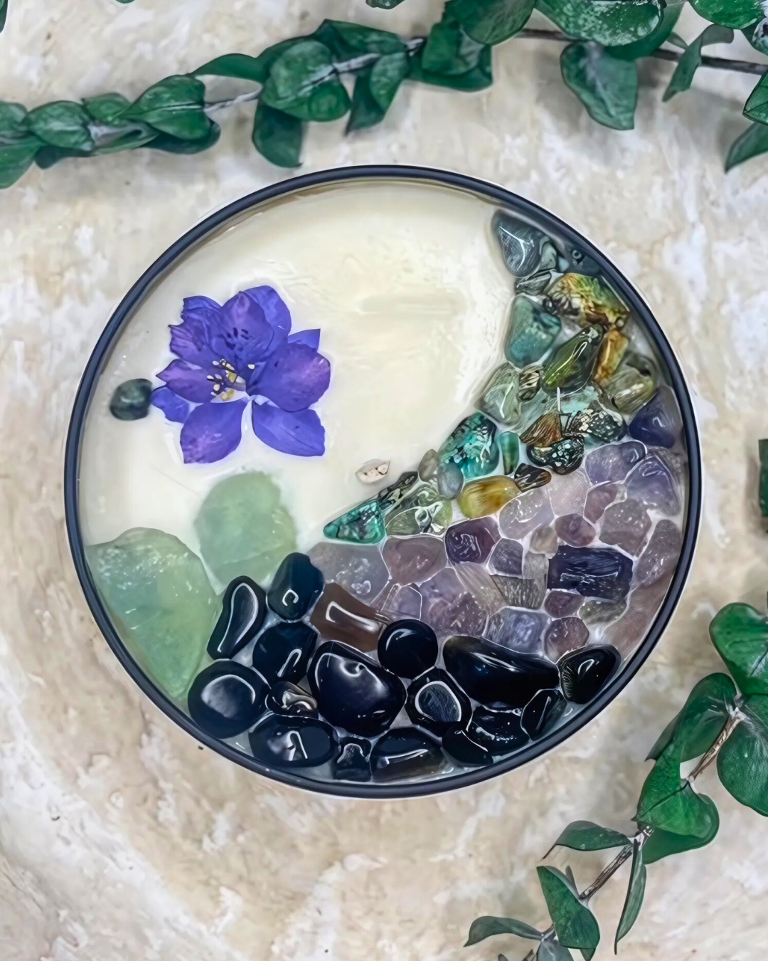 Decorative stone bowl with pebbles and a purple flower, surrounded by green leaves on a textured surface.