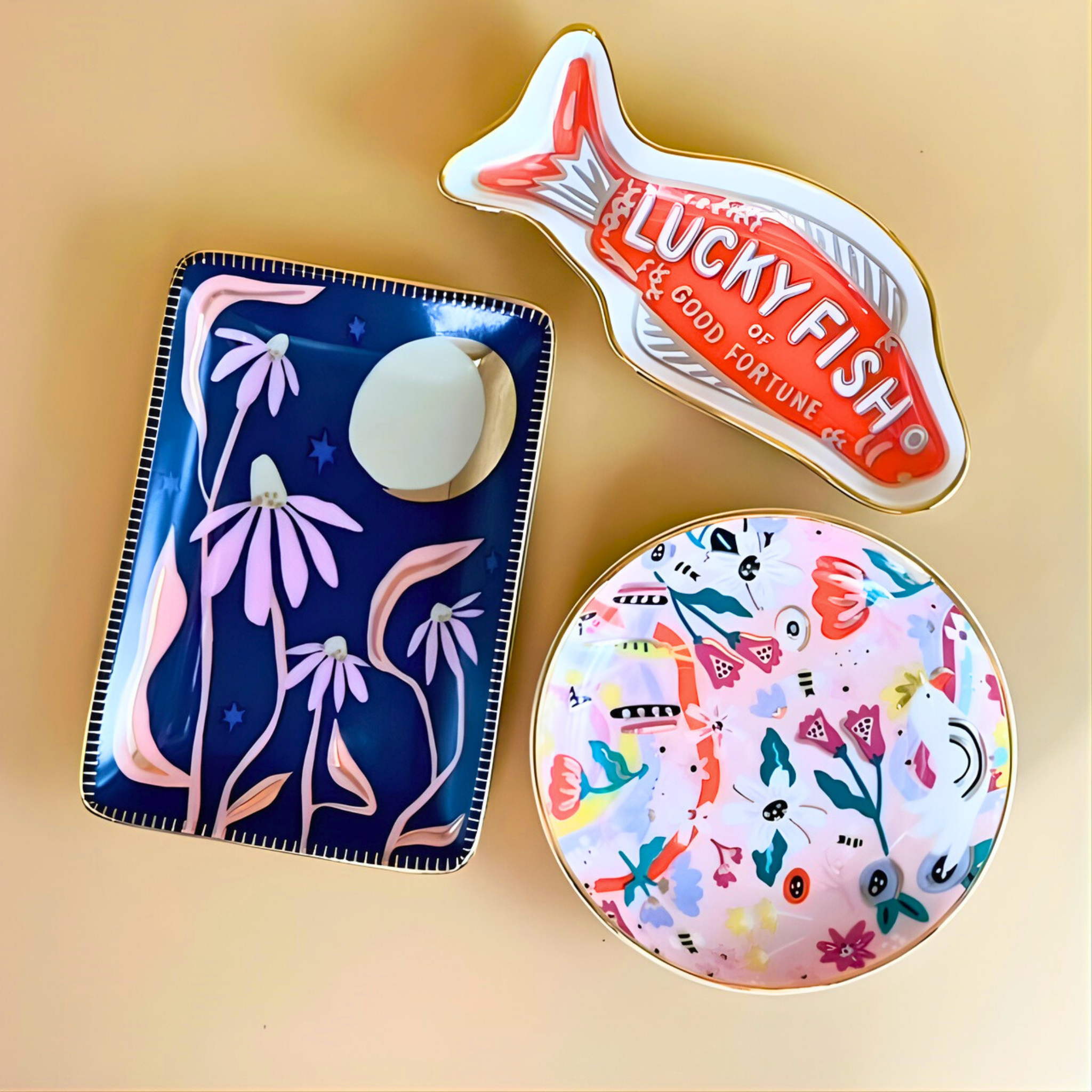Three decorative ring dish on a beige background: a blue pouch with floral design, a round pink coaster with fish and floral patterns, and a rectangular sign with 'Lucky Fish' text.