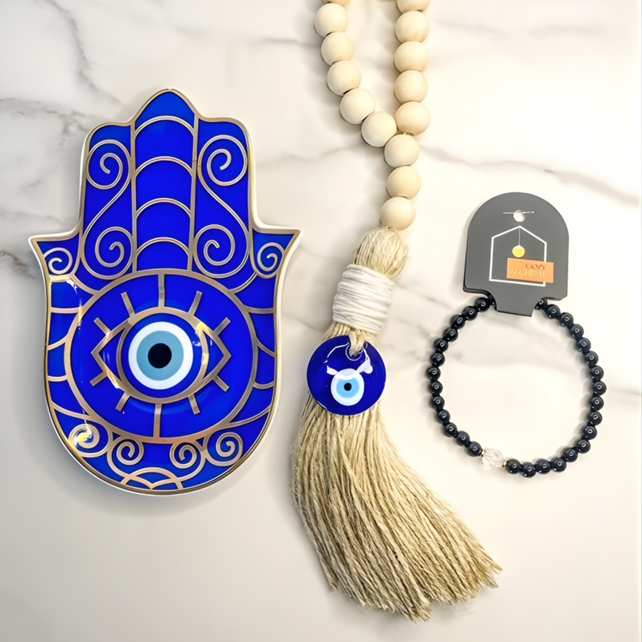Blue Hamsa hand with evil eye, beige tassel, and black beaded bracelet on a marble background