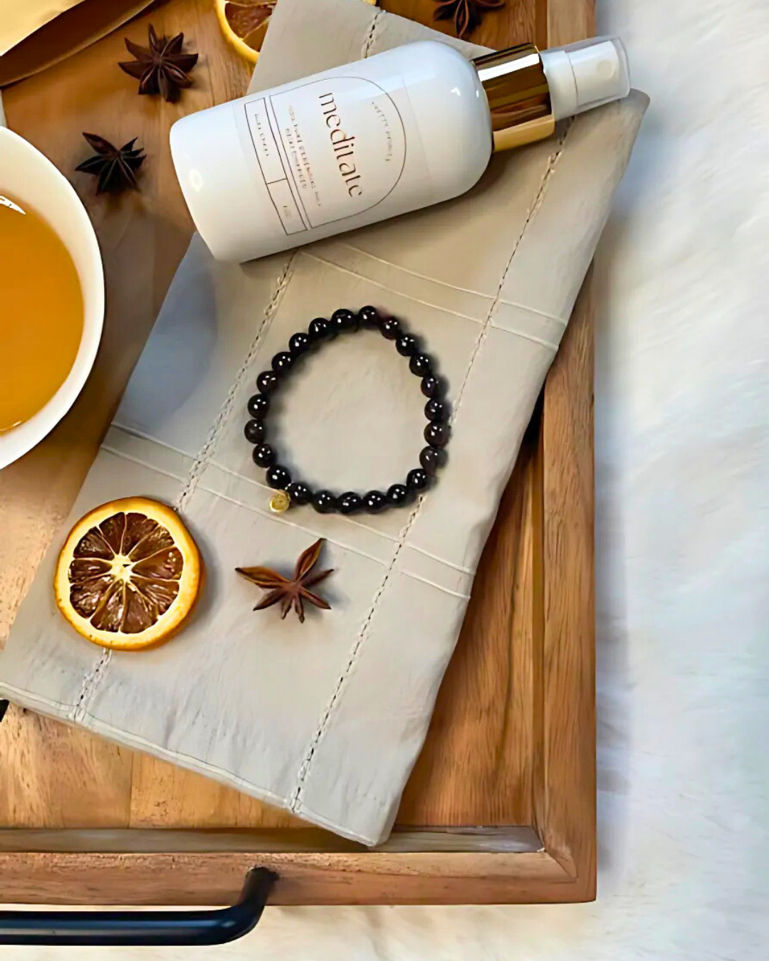 White bottle with gold cap on a wooden tray with a black bracelet, dried orange, and star anise.
