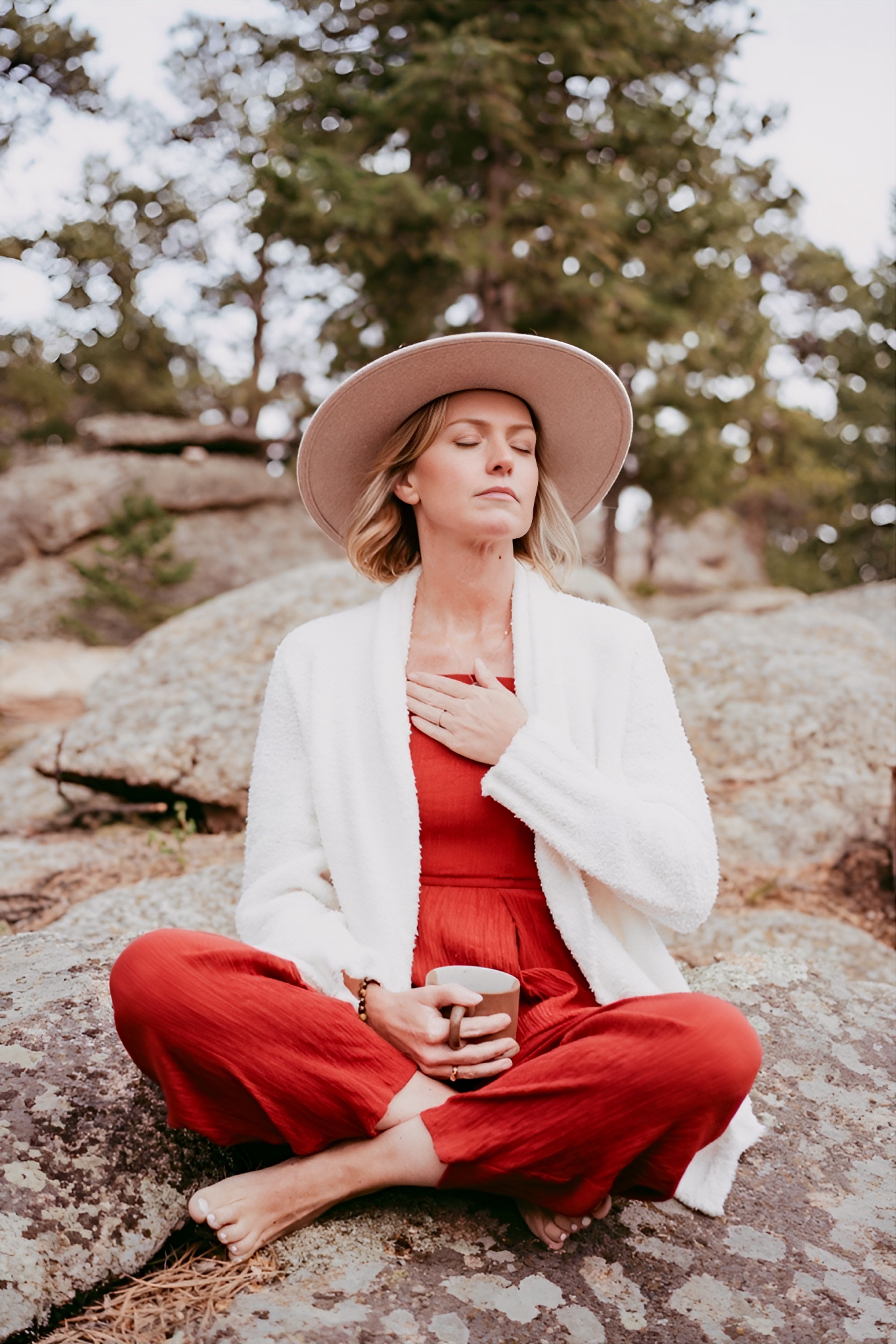Woman in a red outfit and white cardigan sitting on rocks with trees in the background